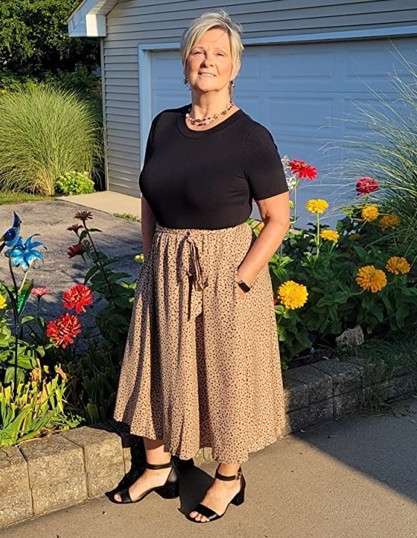 A reviewer stands outside wearing the dress with a black top, black and tan patterned skirt, black sandals, and a necklace