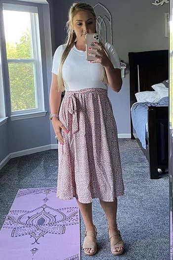 A reviewer takes a mirror selfie wearing the dress with a white top and a pink and white patterned skirt and sandals