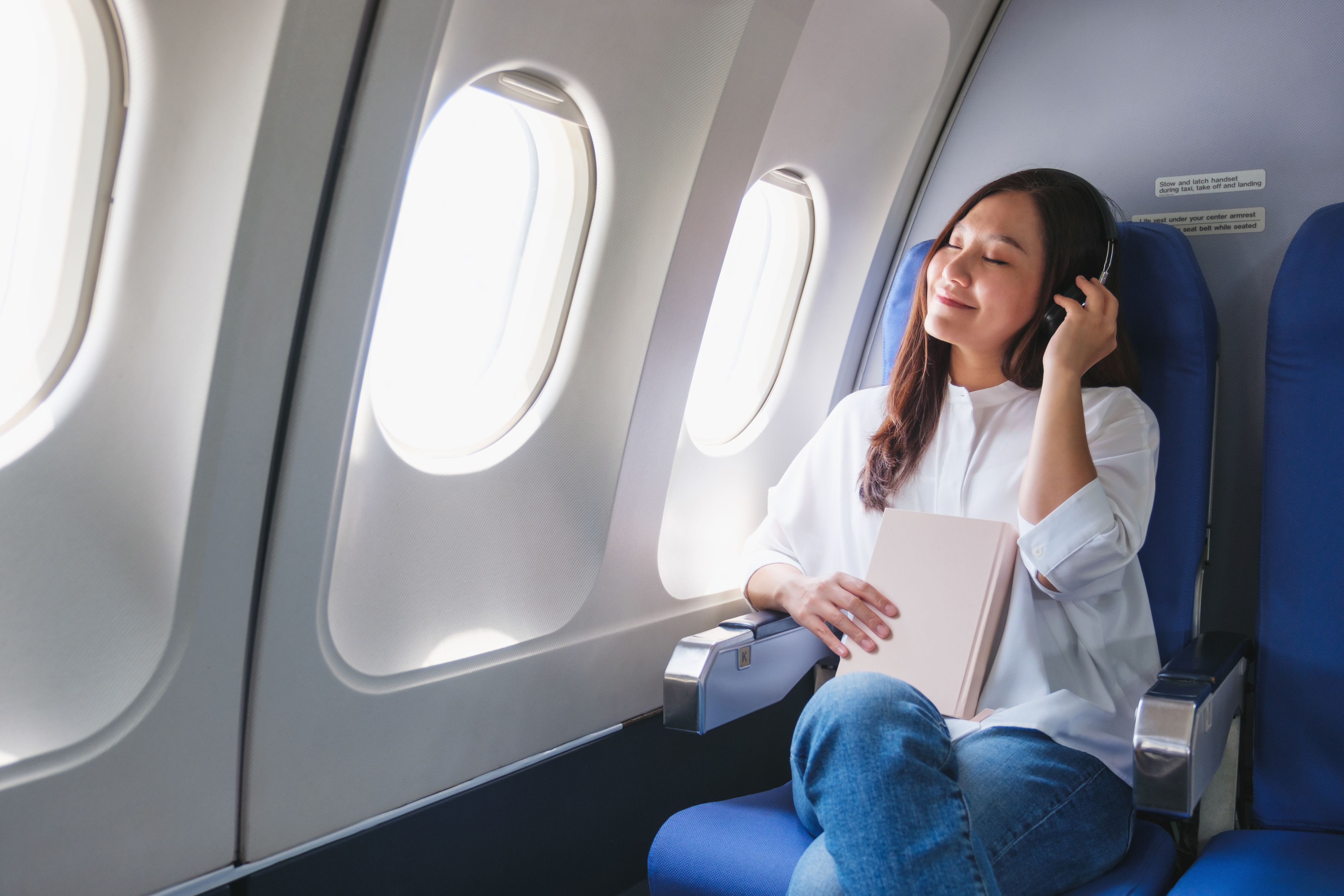 A woman sits comfortably in an airplane seat by the window, enjoying music with headphones and holding a book