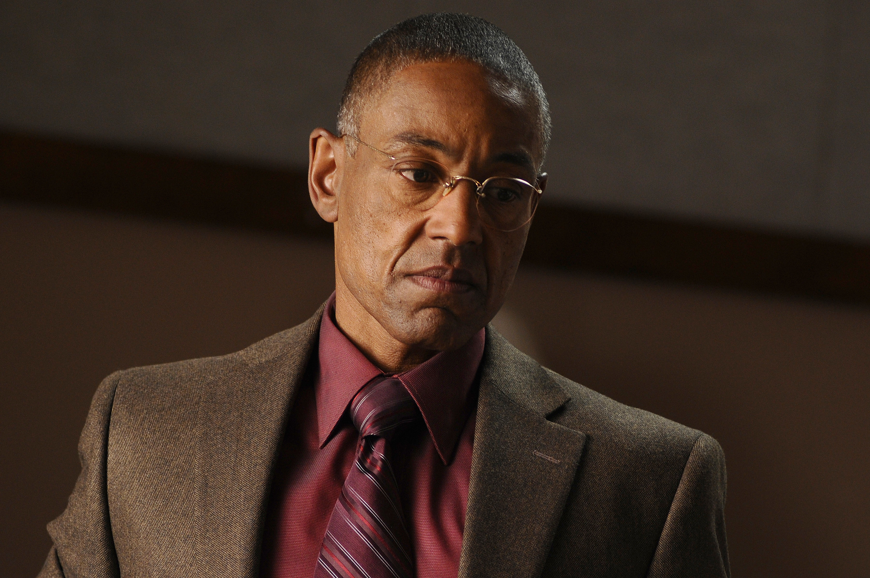 Giancarlo Esposito wearing a suit jacket with a dress shirt and tie, looking down pensively in a scene from "Breaking Bad"