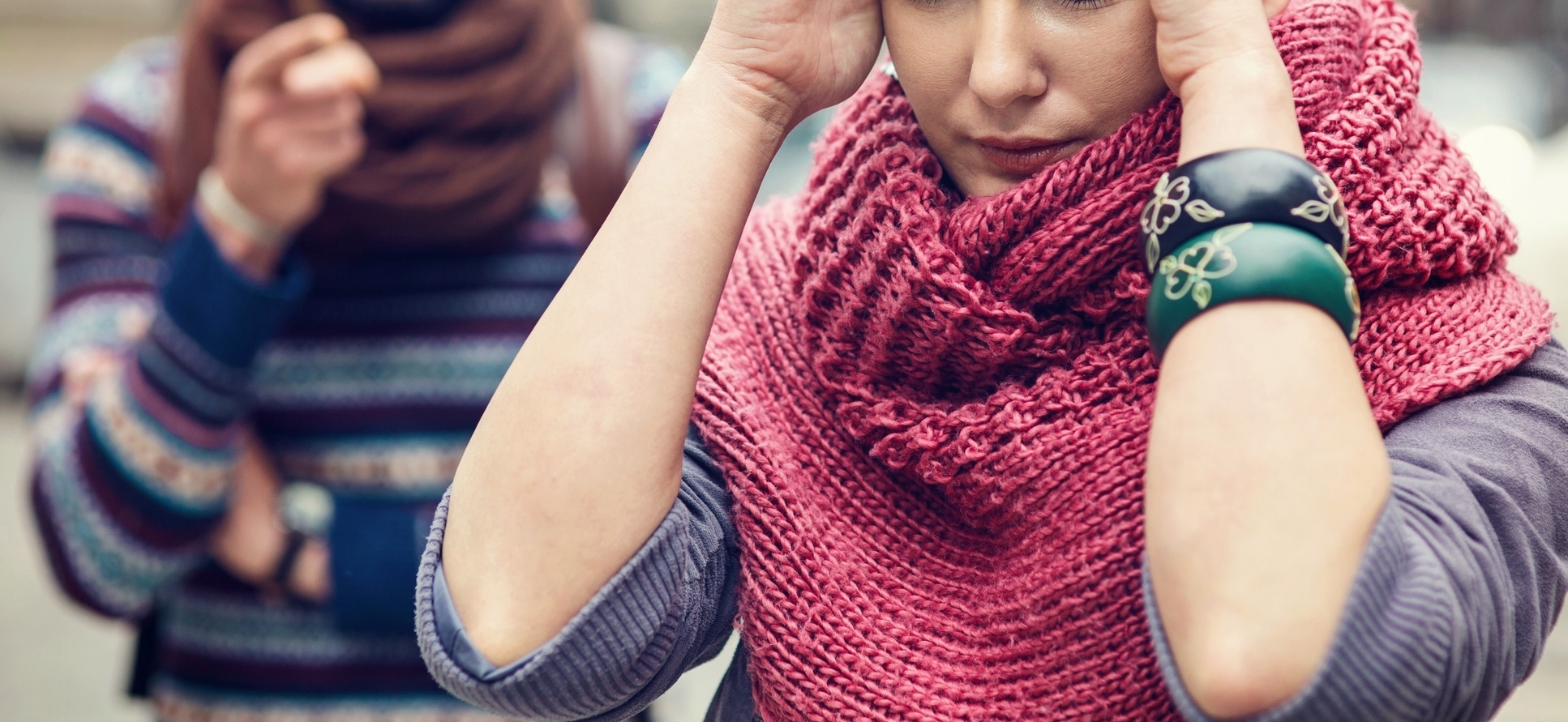 A person in a knitted shawl looks distressed, while another person with folded arms stands in the background