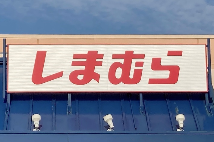 Storefront with a sign displaying Japanese text reading “Shimamura”. The building has a blue facade and a clear sky above