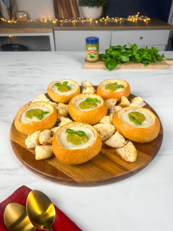 Bread bowls filled with soup, garnished with fresh herbs, surrounded by croutons on a wooden platter. Pesto jar and fresh greens are in the background on a kitchen counter