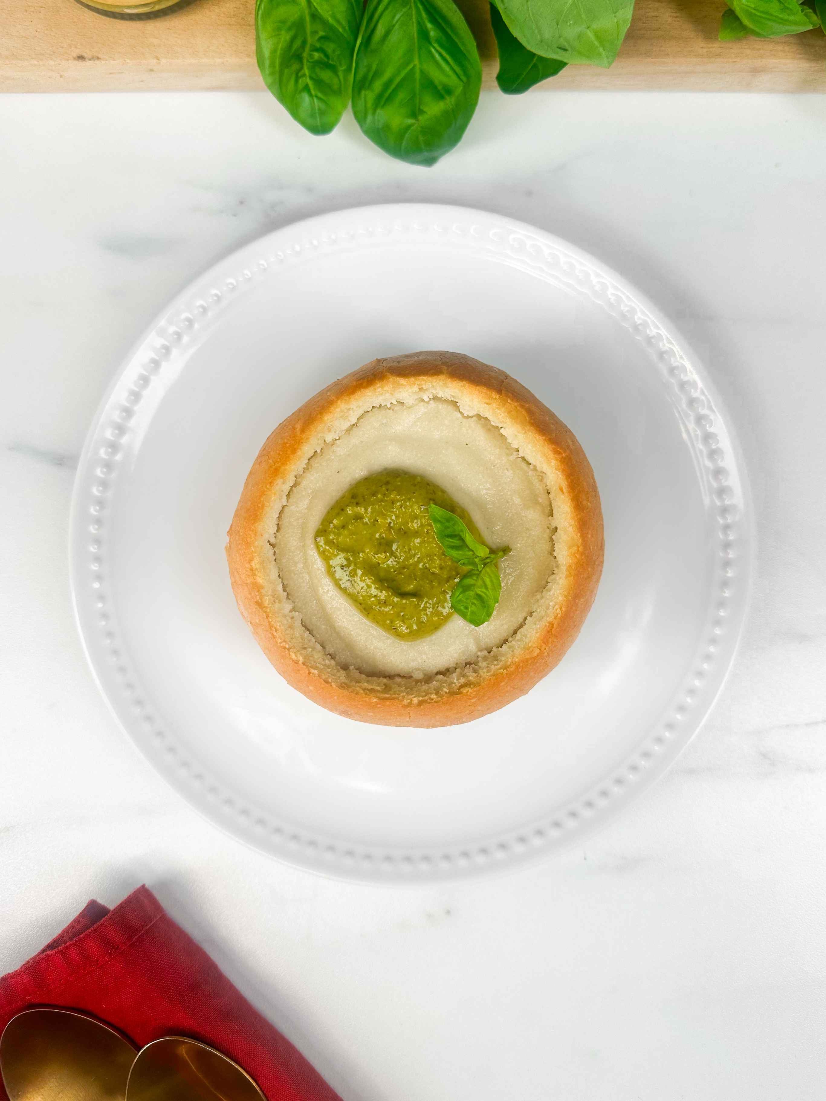 A bowl of creamy soup served in a bread bowl, garnished with a dollop of pesto and basil leaves, with some fresh basil in the background