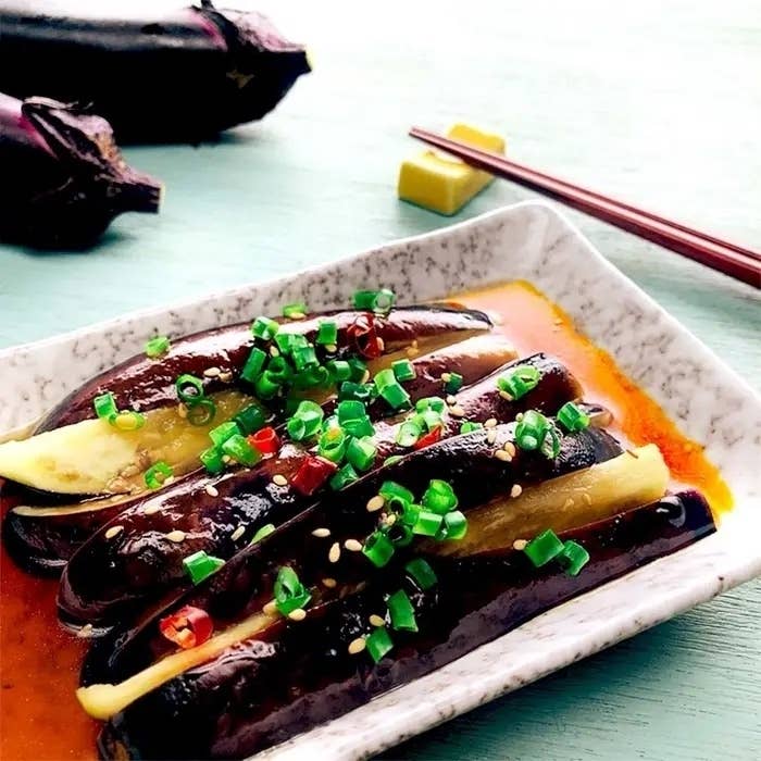 白い皿に並べられた細長いナスの料理。ナスの上には刻んだネギと唐辛子が載っている。後ろには箸と他のナスも見える。