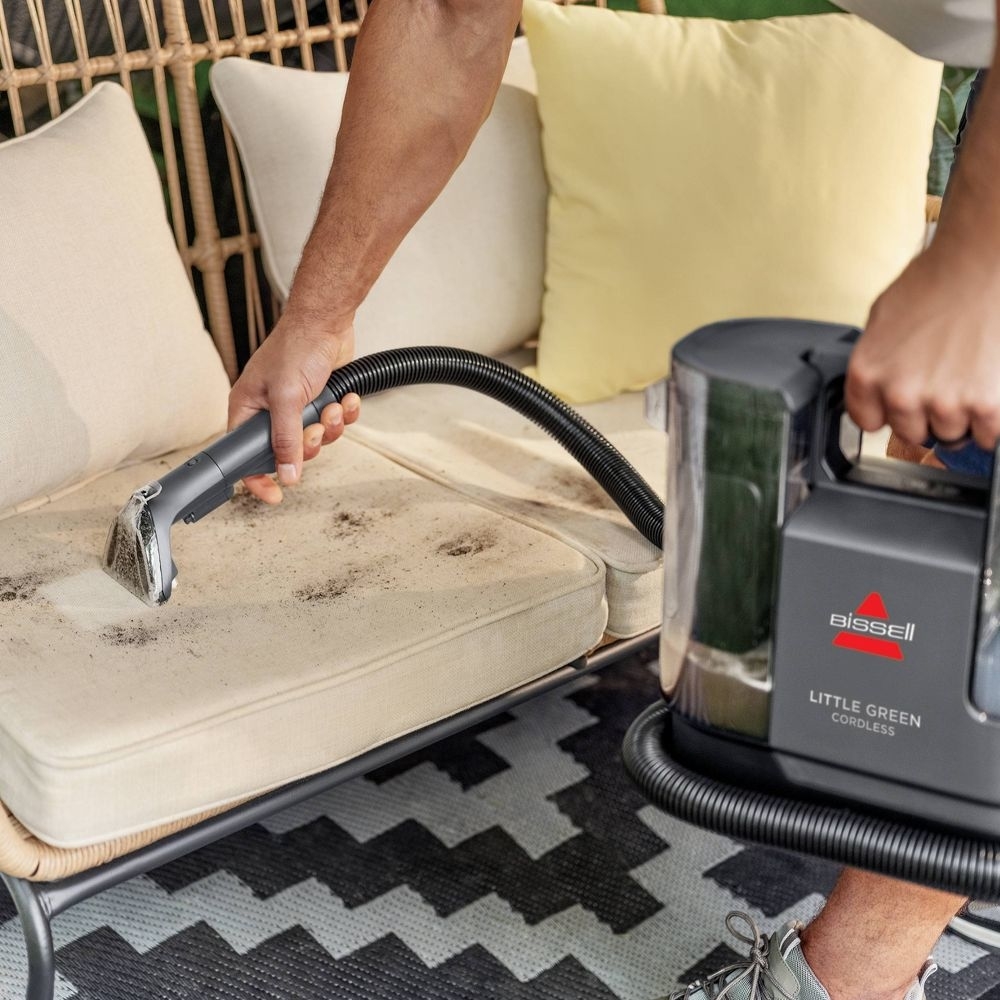 Person using a Bissell Little Green cordless cleaner to remove dirt from a light-colored couch cushion in an outdoor setting