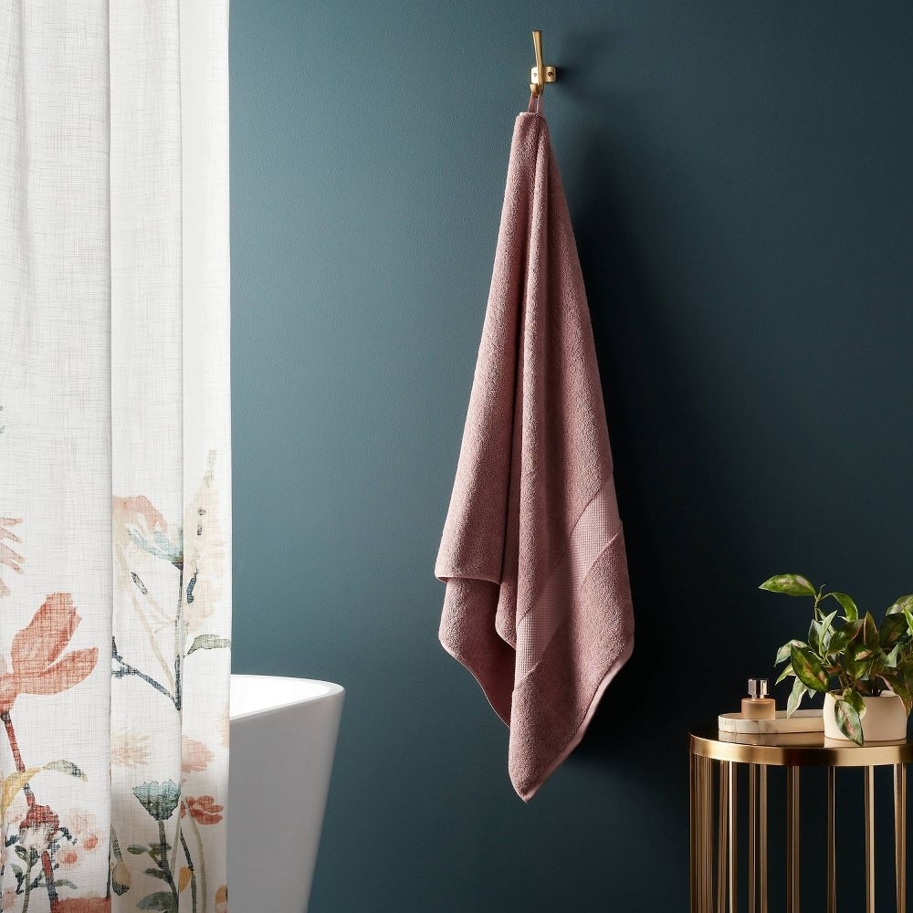 A pink towel hangs on a gold hook next to a floral shower curtain and a small, round table with a potted plant