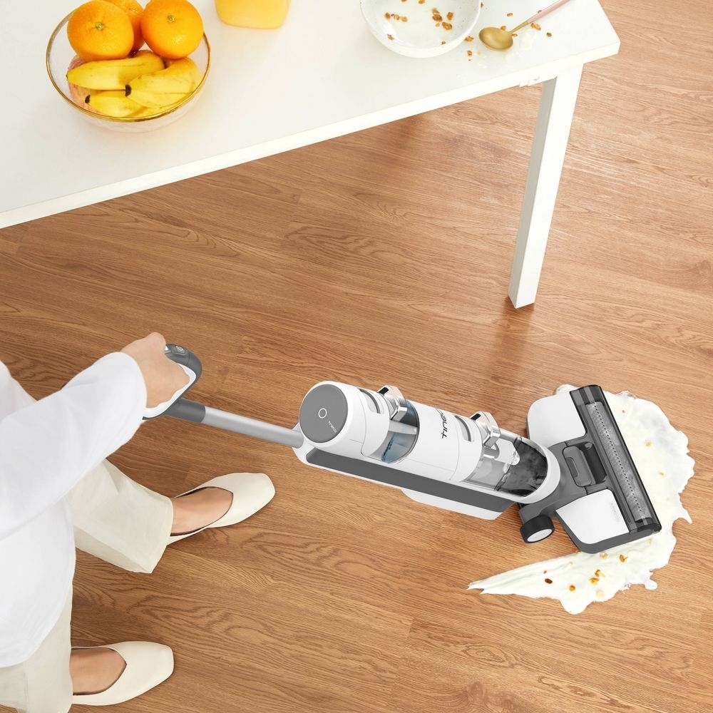 Person cleaning spilled milk and crumbs on a wooden floor with a lightweight, cordless vacuum near a table with fruit