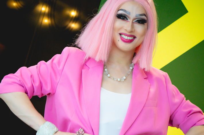 人物がカメラにポーズをとっている。ピンクのウィッグとピンクのブレザーを着用し、大きな笑顔を見せている。