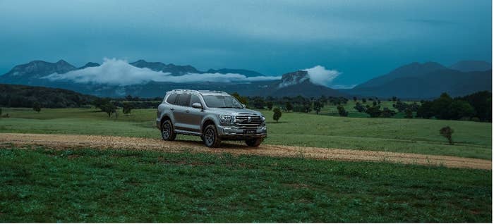 An SUV is parked on a dirt road in a lush green open field with mountains and clouds in the background. No people are visible