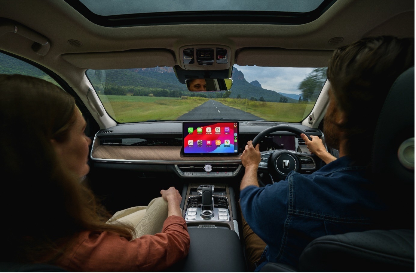 A woman and man are in a car driving on a scenic road. The car’s dashboard displays navigation and other apps