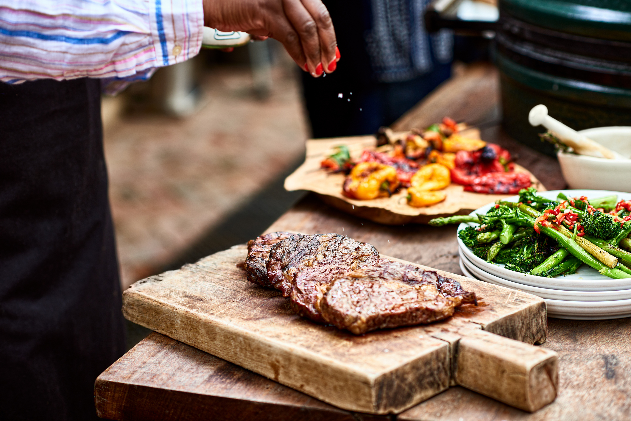 A person seasons a cooked steak on a wooden cutting board. In the background, grilled vegetables and a bowl of greens are on display