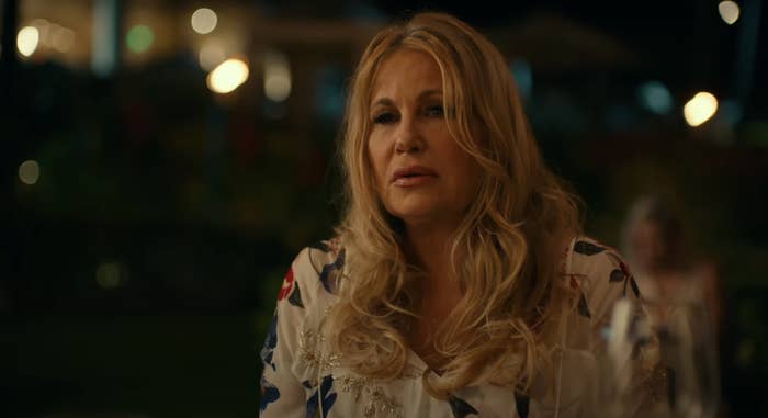 Jennifer Coolidge in an elegant, floral outfit, seated at a formal event, appearing thoughtful, in a scene from White Lotus