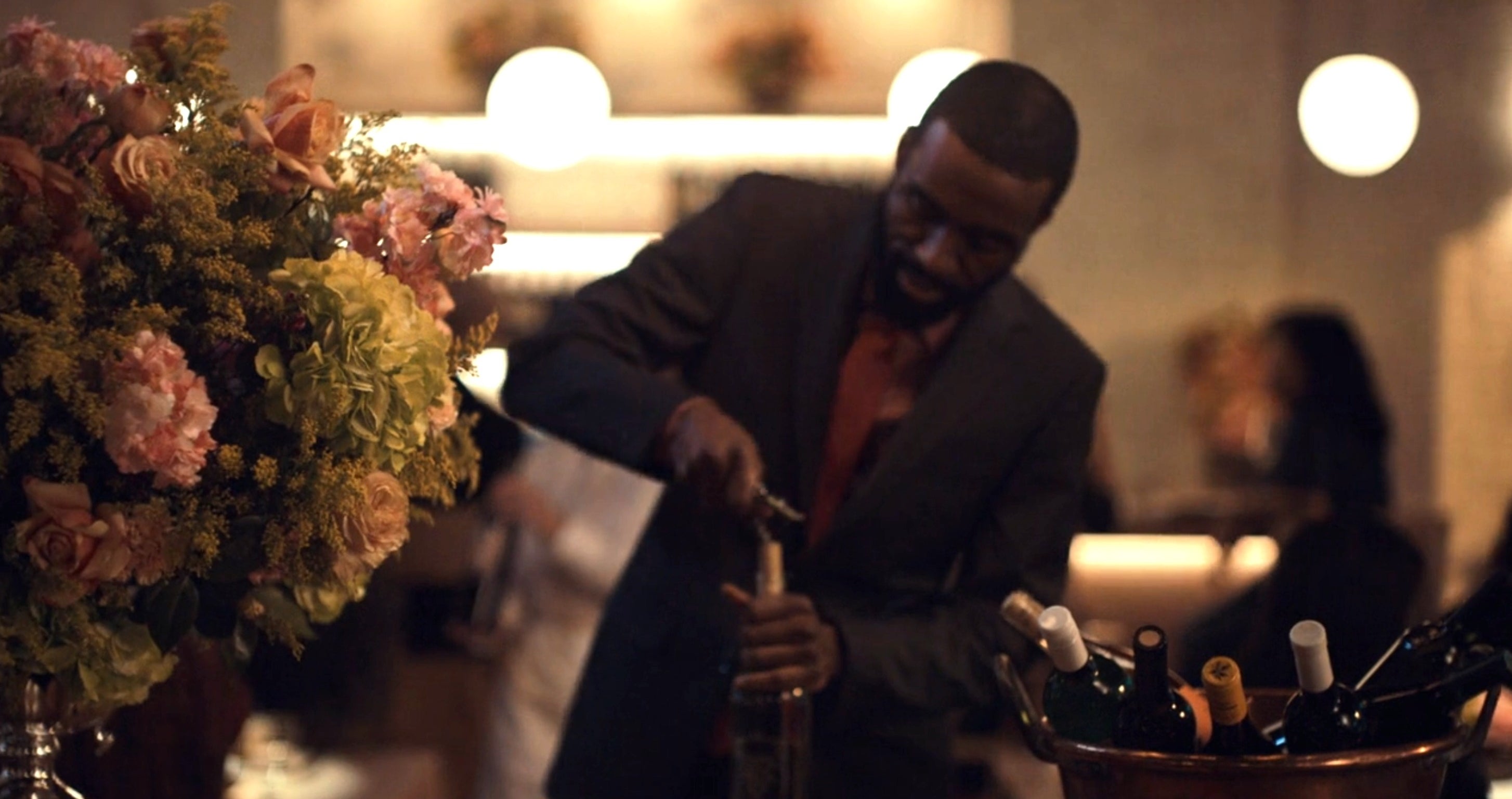 A man in formal attire opens a bottle of wine at an upscale event, with flowers and other guests in the background
