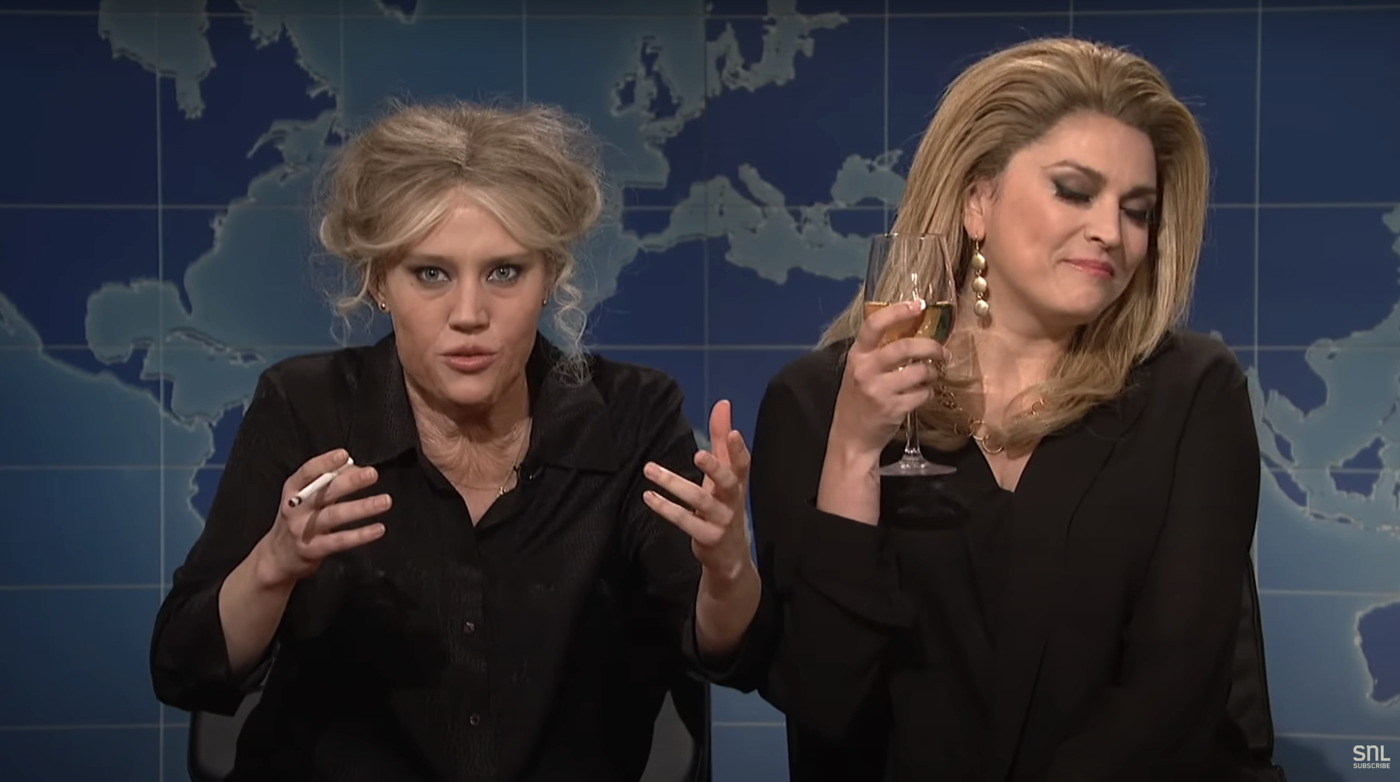 Kate McKinnon and Cecily Strong in a news show parody scene, with Kate speaking animatedly and Cecily holding a wine glass