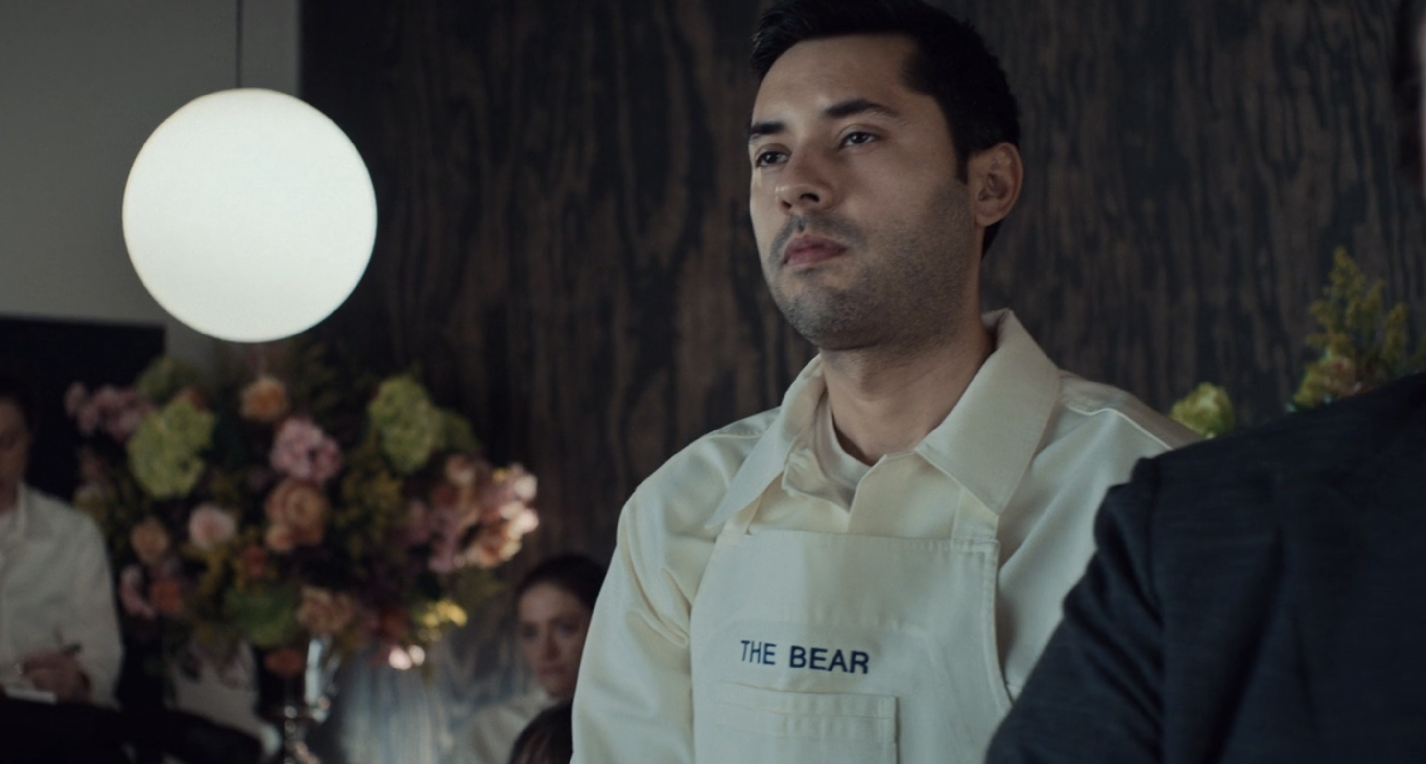 A man wearing a shirt and apron labeled &quot;The Bear&quot; stands in a professional setting with flowers in the background