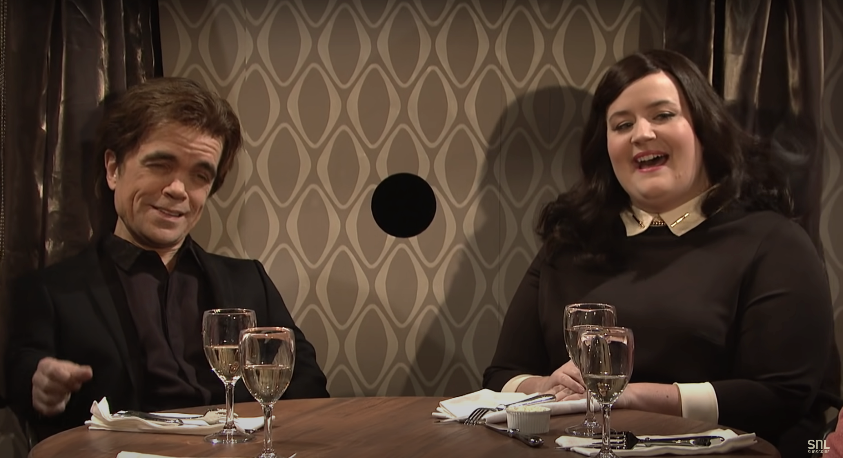 Peter Dinklage and Aidy Bryant sitting at a round table with wine glasses, engaged in conversation on a stage set resembling a restaurant