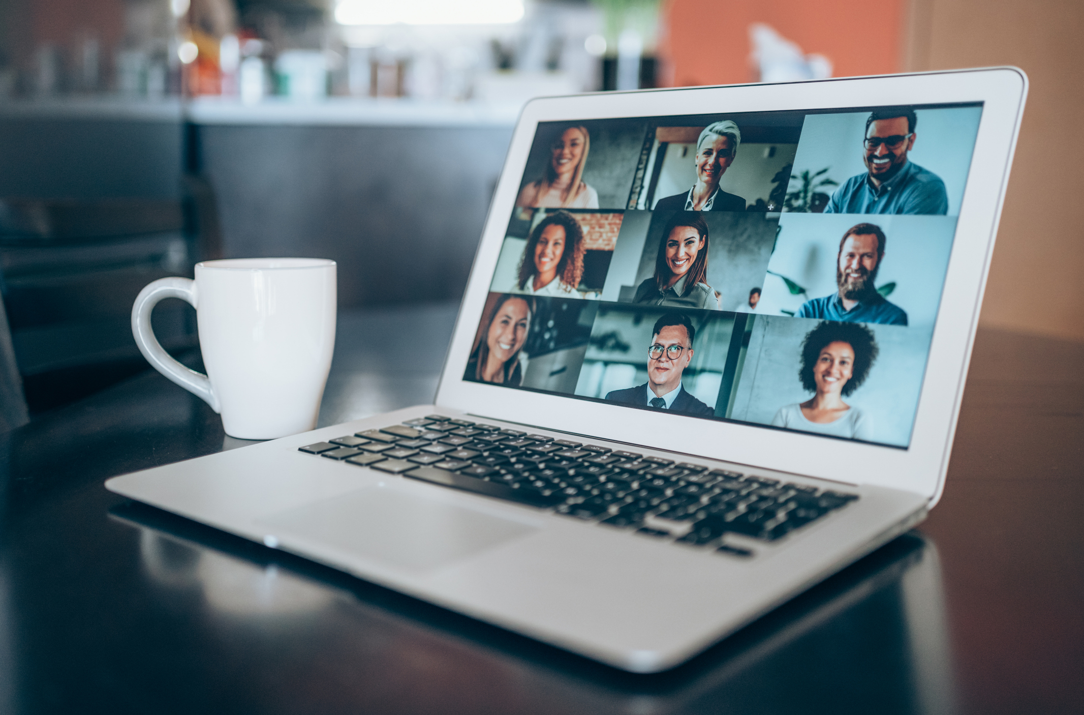 A laptop on a desk displays a video conference call with ten participants. A coffee mug is to the left of the laptop