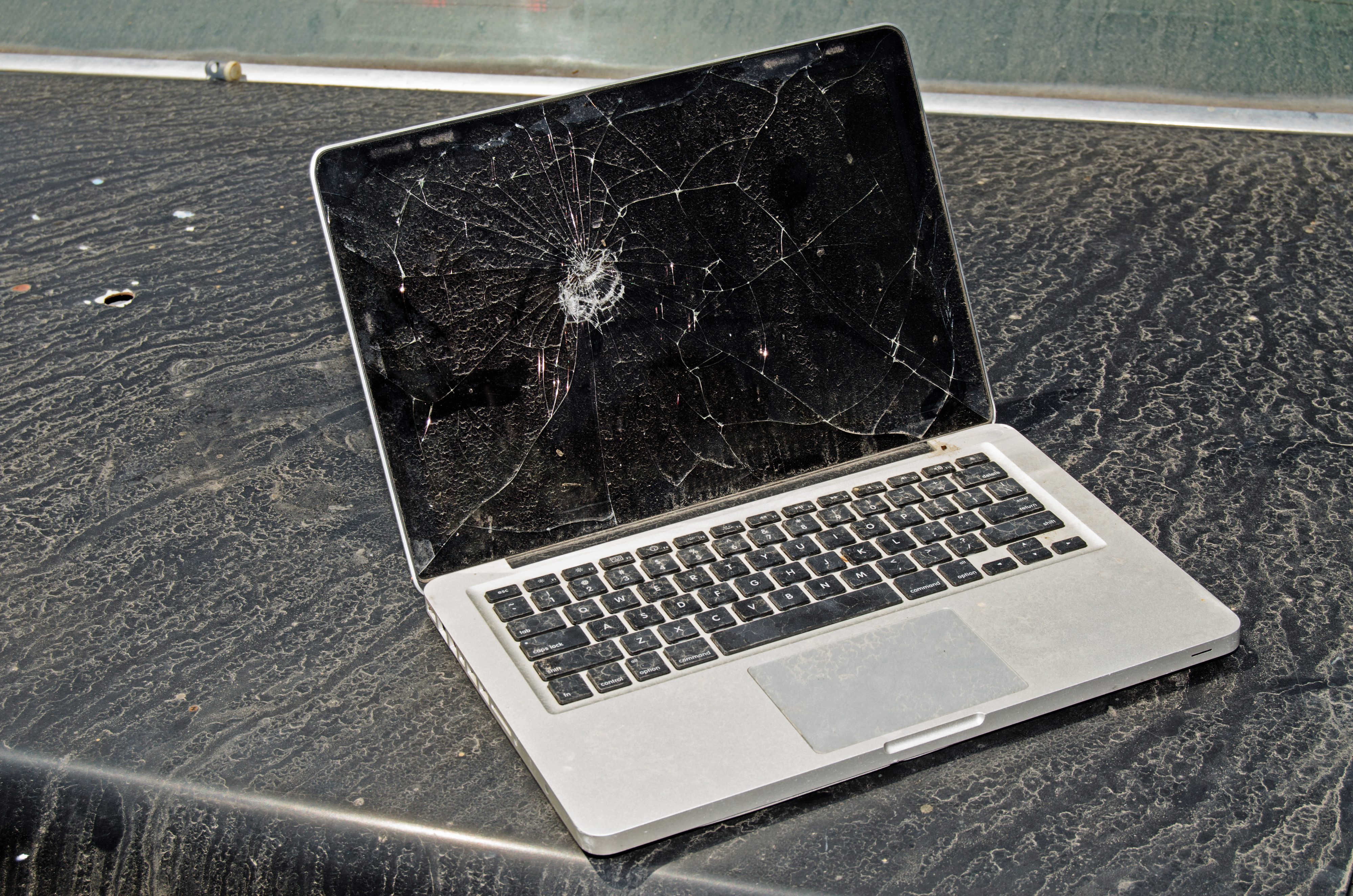 A laptop with a shattered screen is placed on a dusty surface. The device appears significantly damaged and non-functional