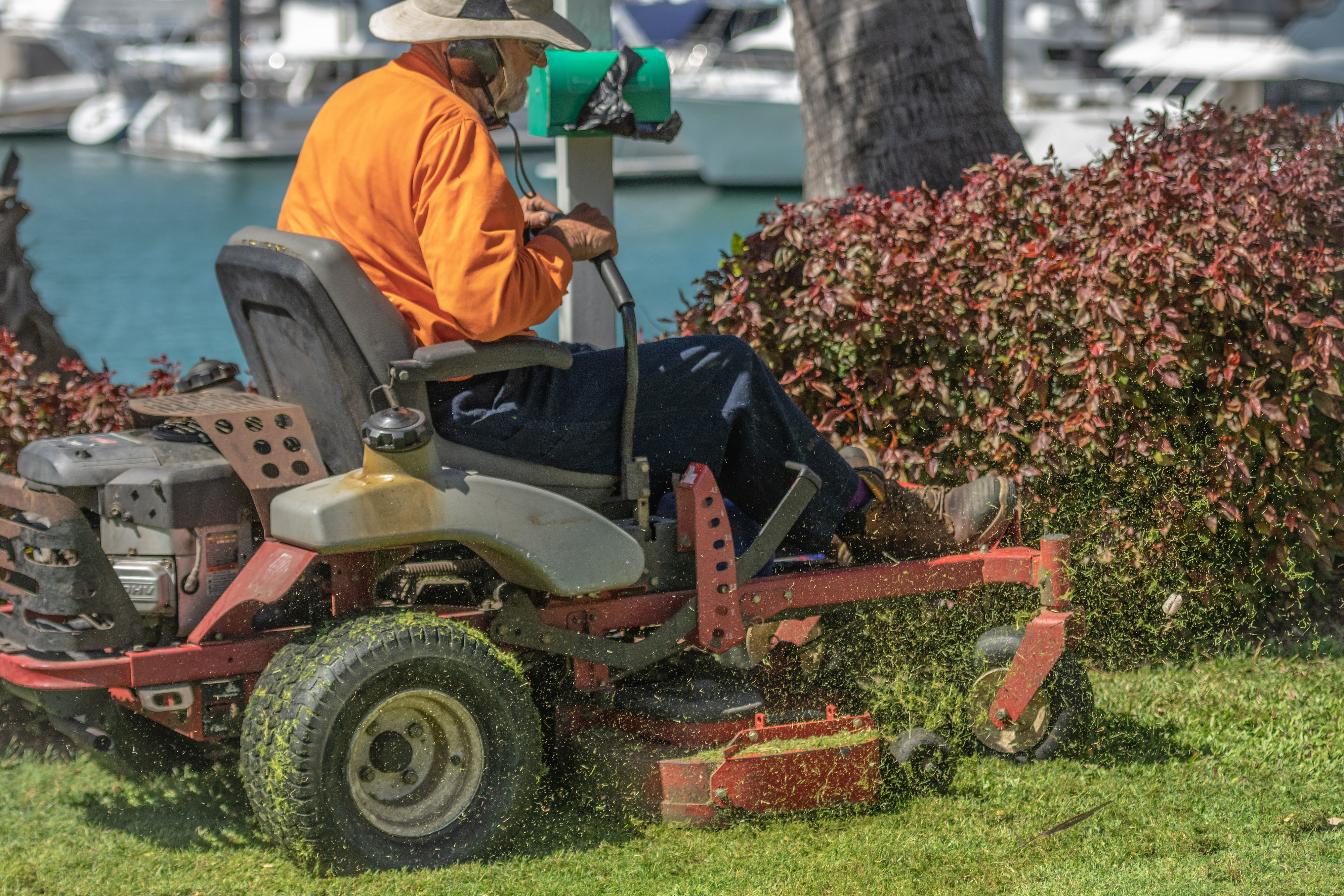 A person wearing an orange long-sleeved shirt operates a riding lawnmower next to hedges with boats and water in the background