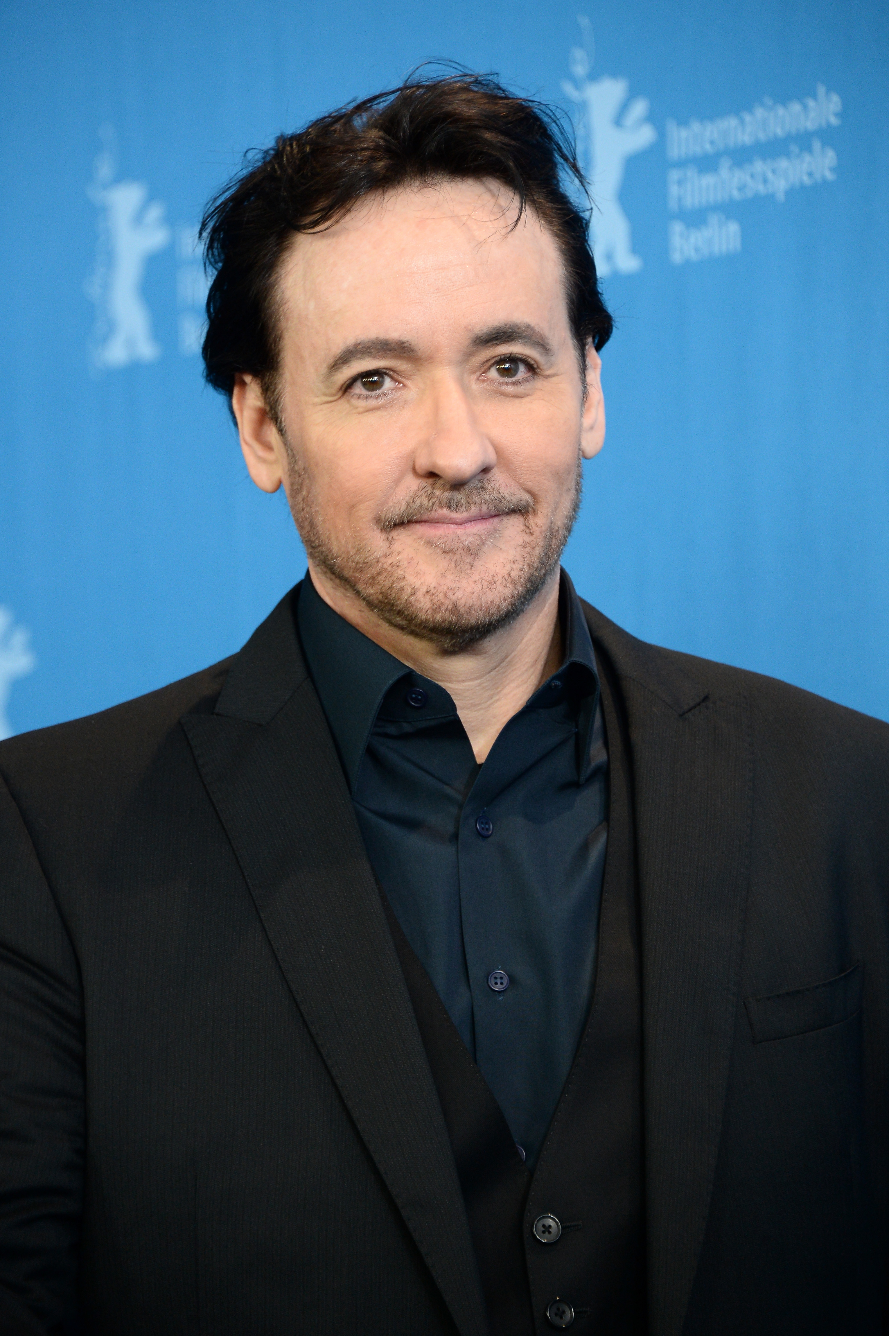 John Cusack in a dark suit and black shirt poses in front of a backdrop at the Berlin International Film Festival
