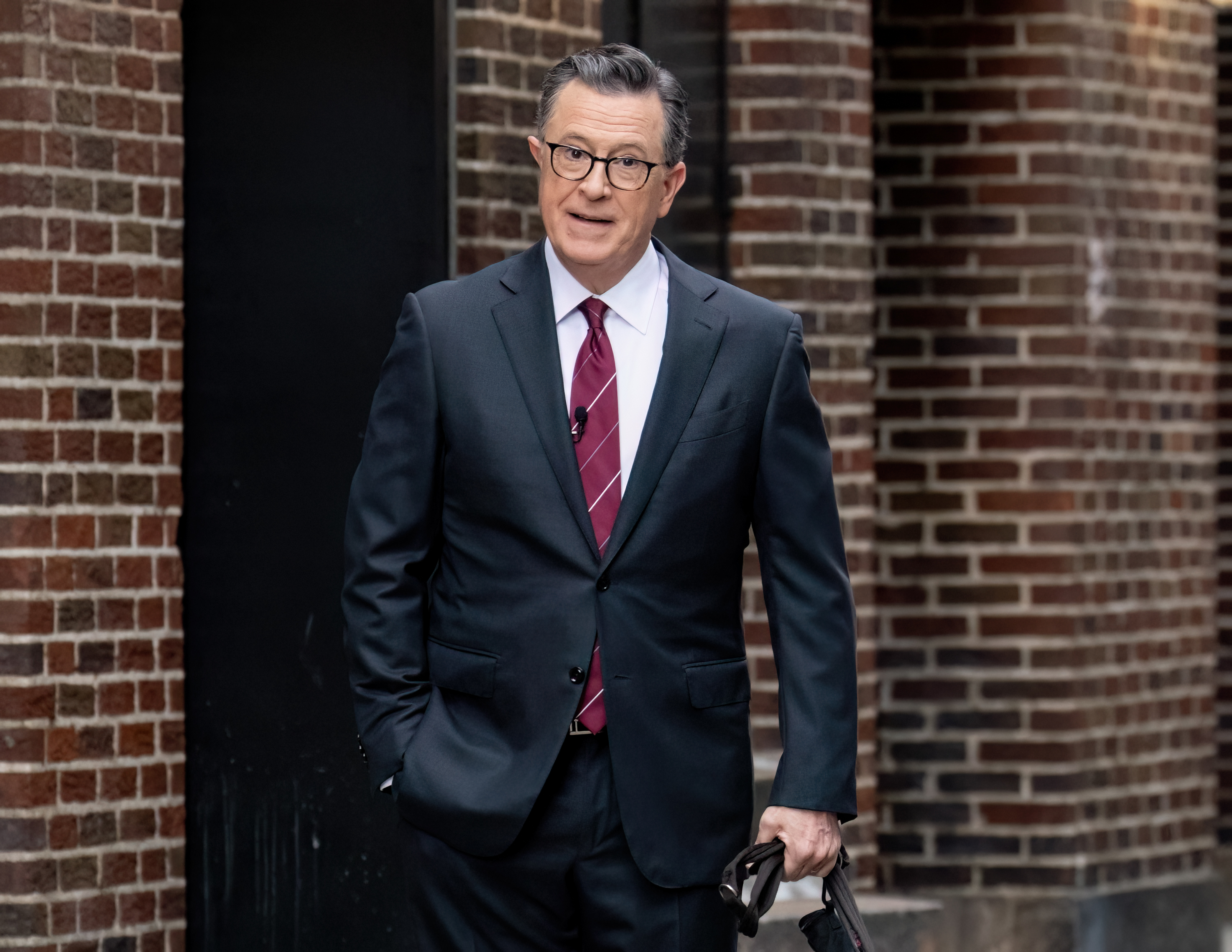 Stephen Colbert is walking outdoors, dressed in a dark suit, white shirt, and striped tie, holding glasses in one hand, with his other hand in his pocket