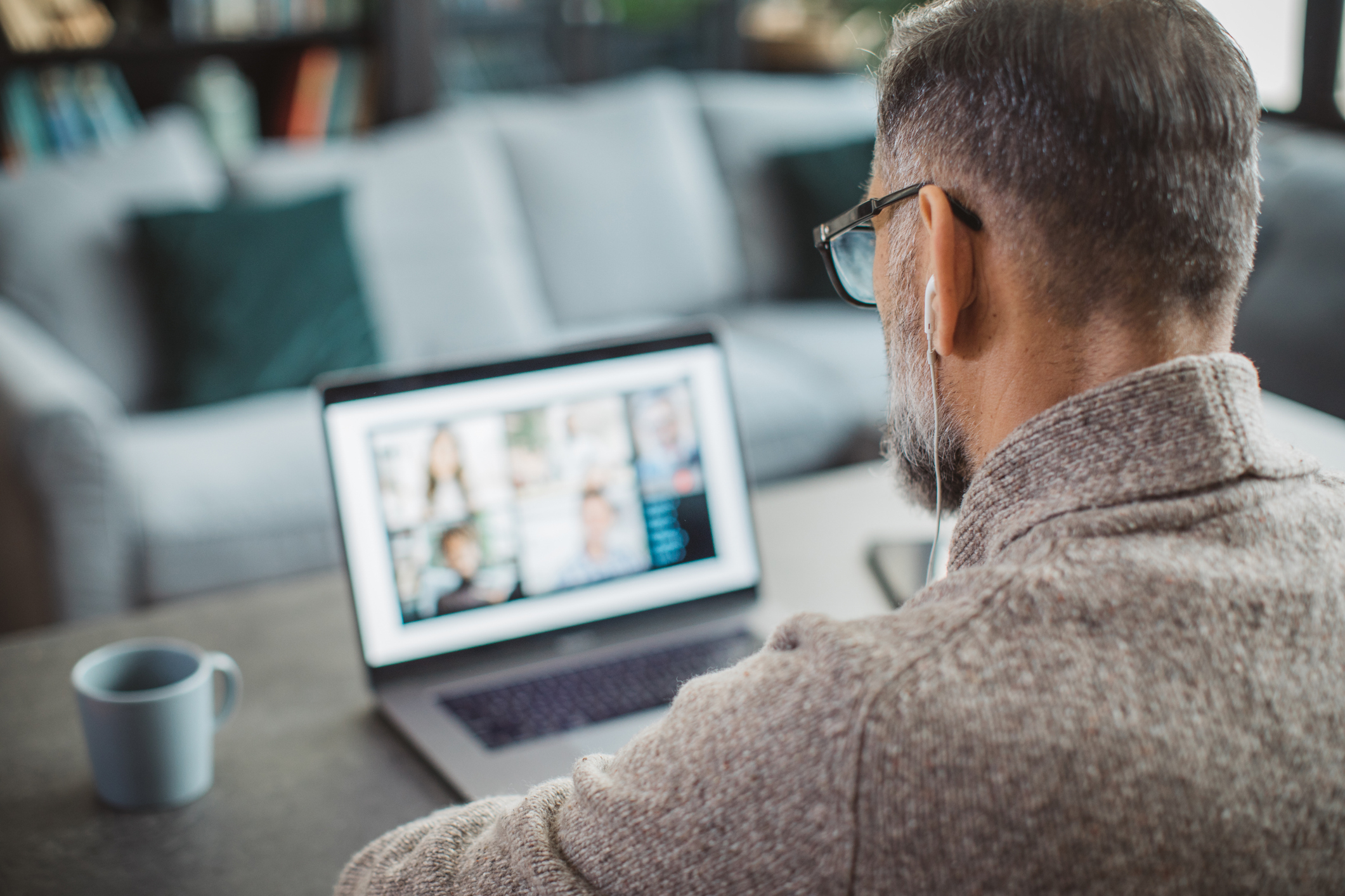 A man with glasses and earbuds works on a laptop at home. The screen shows multiple small images during a video conference