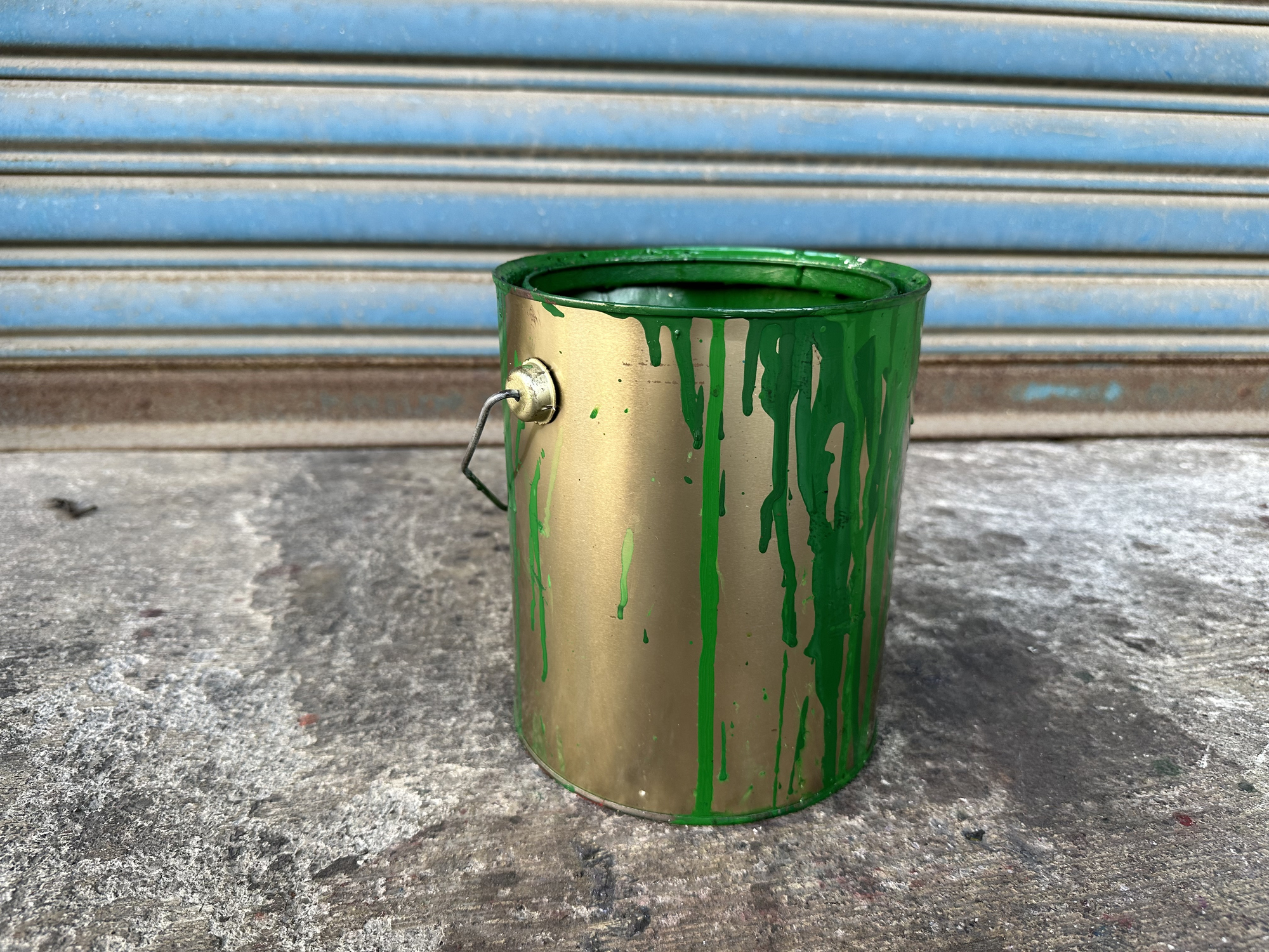 A paint can with spilled green paint sits on the ground in front of a metal shutter