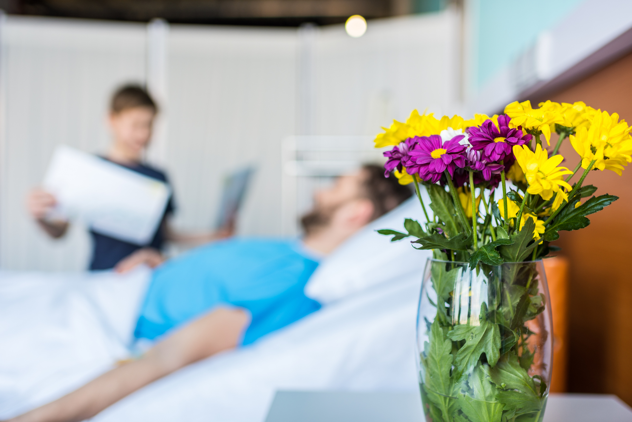 A vase of flowers on a table with a person lying in a hospital bed and another person standing and holding a document in the background