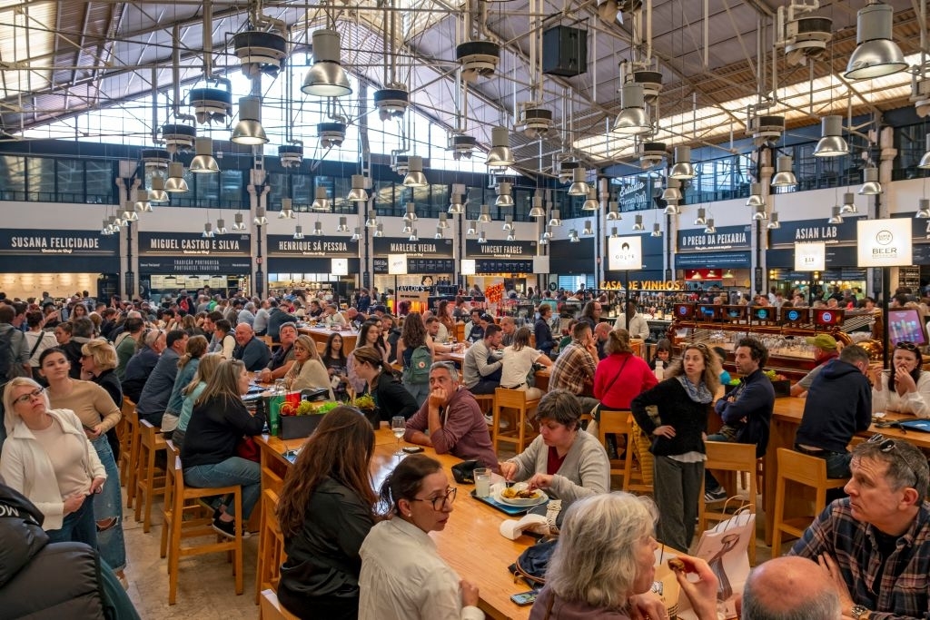 People dining and socializing at a busy indoor marketplace with various food stalls. The atmosphere is lively, with patrons seated at communal tables