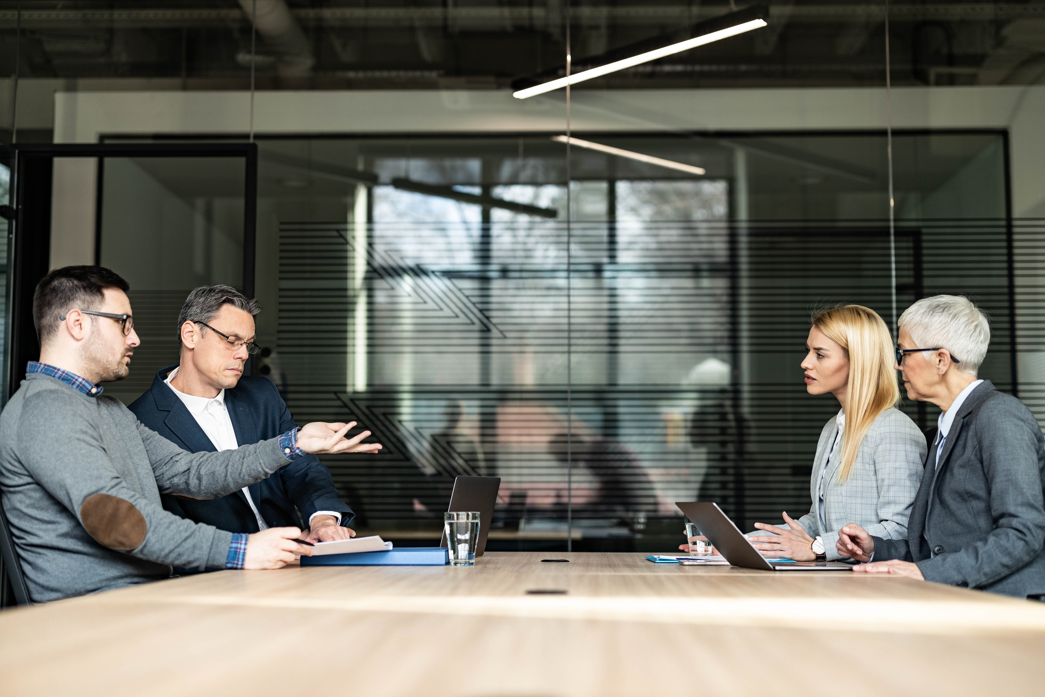 A business meeting with four people in a modern office. Two men on the left are gesturing, while two women on the right listen attentively