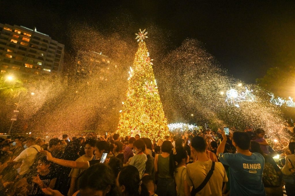 A large crowd celebrates around a tall, illuminated Christmas tree in a bustling city square at night, with festive lights and artificial snow enhancing the atmosphere