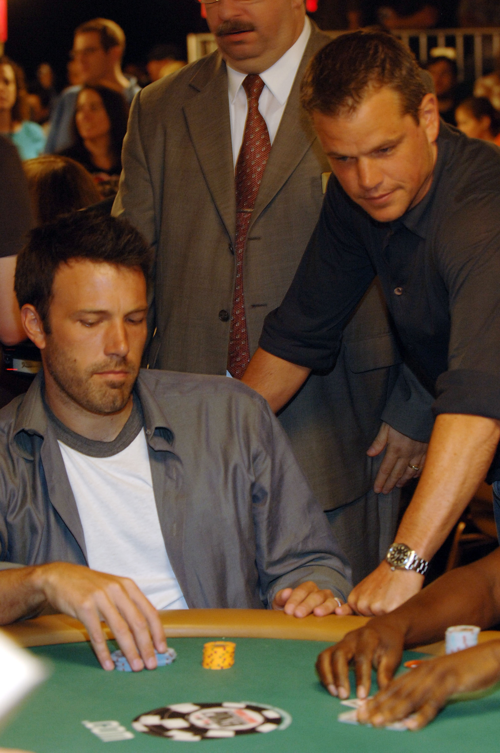 Ben Affleck and Matt Damon at a poker table during a tournament, concentrating on their cards. Affleck is seated, and Damon is standing behind him