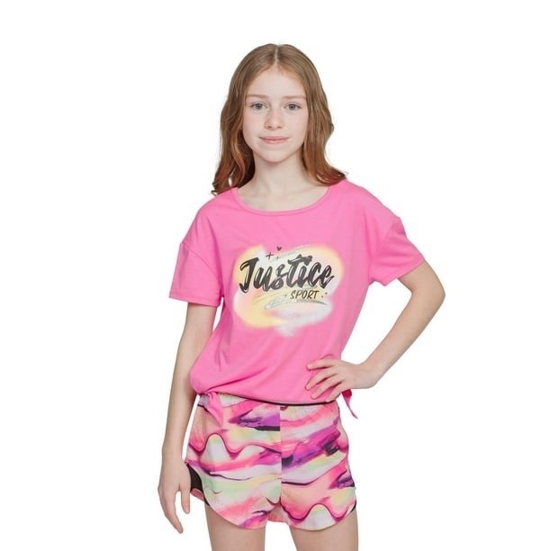 A girl poses in a Justice Sport T-shirt and vibrant abstract-patterned shorts, smiling while standing with one hand on her hip