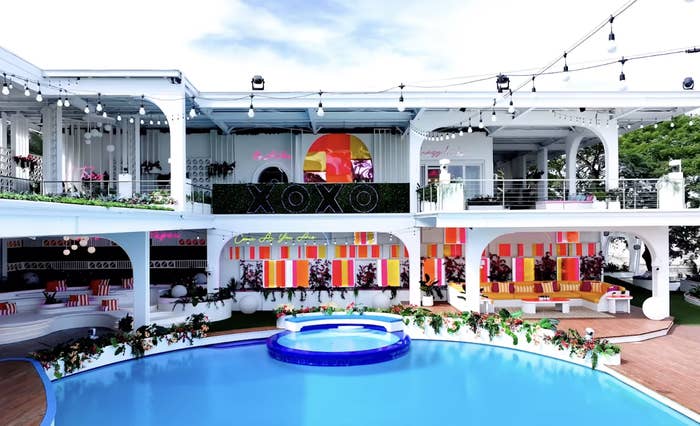 Luxurious villa with a pool, two-story structure, string lights, colorful interior décor, and greenery. "XOXO" sign visible on upper level