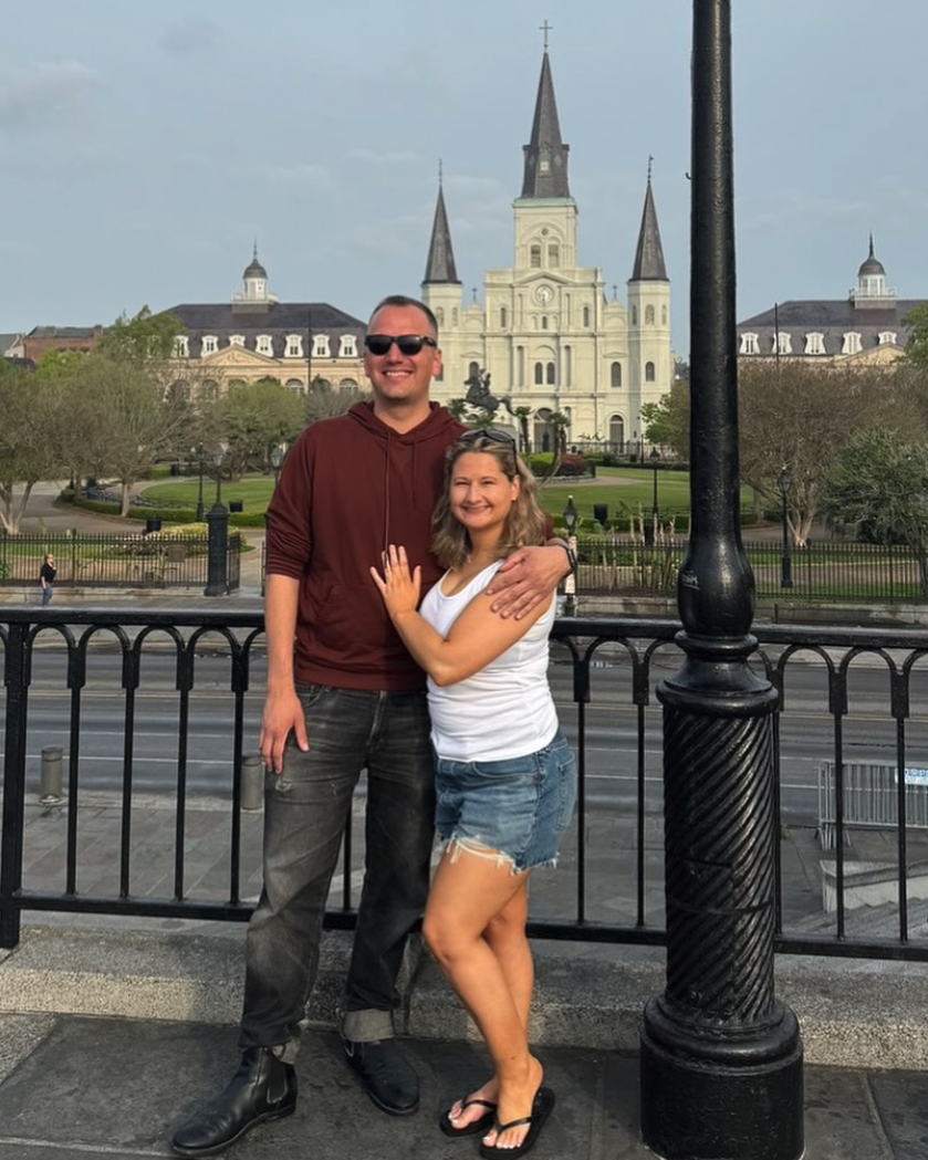 A man (dark sunglasses, dark red hoodie) and woman (denim shorts, white sleeveless top) pose in front of St. Louis Cathedral in New Orleans, smiling