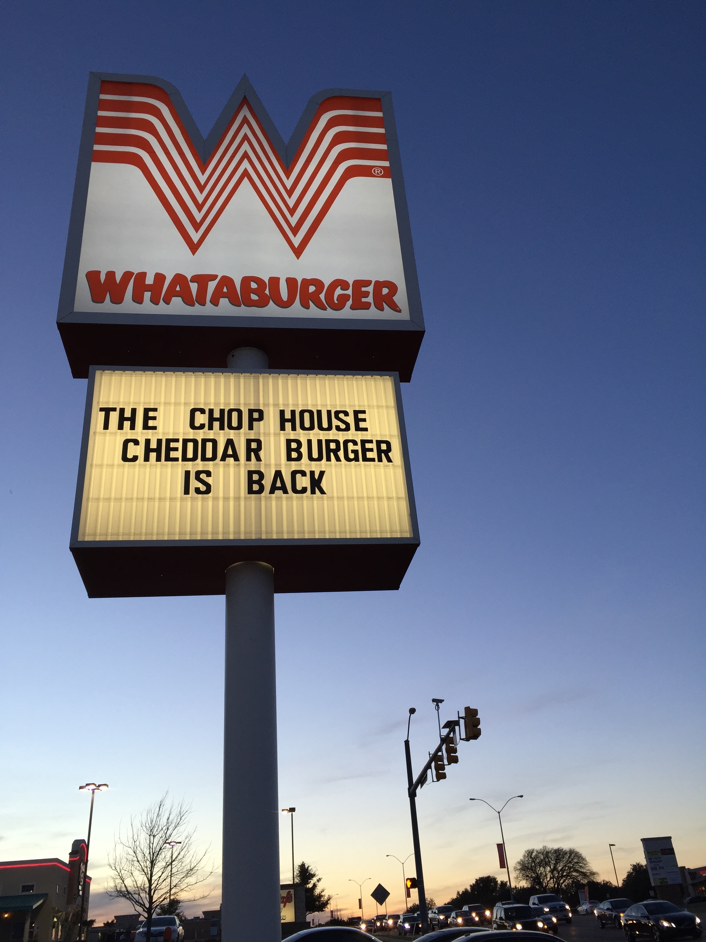 Whataburger sign at dusk announcing "THE CHOP HOUSE CHEDDAR BURGER IS BACK" at a street intersection
