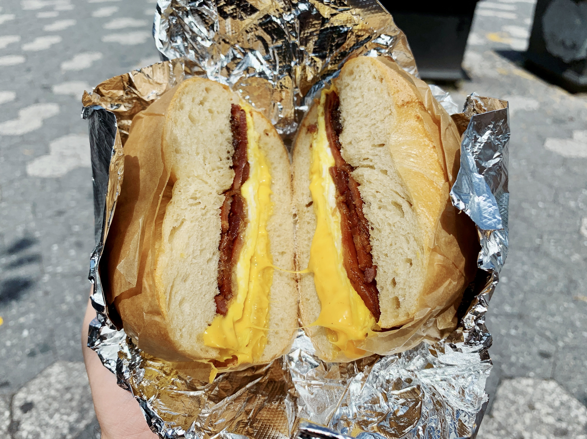 A close-up of a cut breakfast sandwich with eggs, bacon, and cheese, wrapped in foil. The sandwich is held outdoors on a sunlit sidewalk