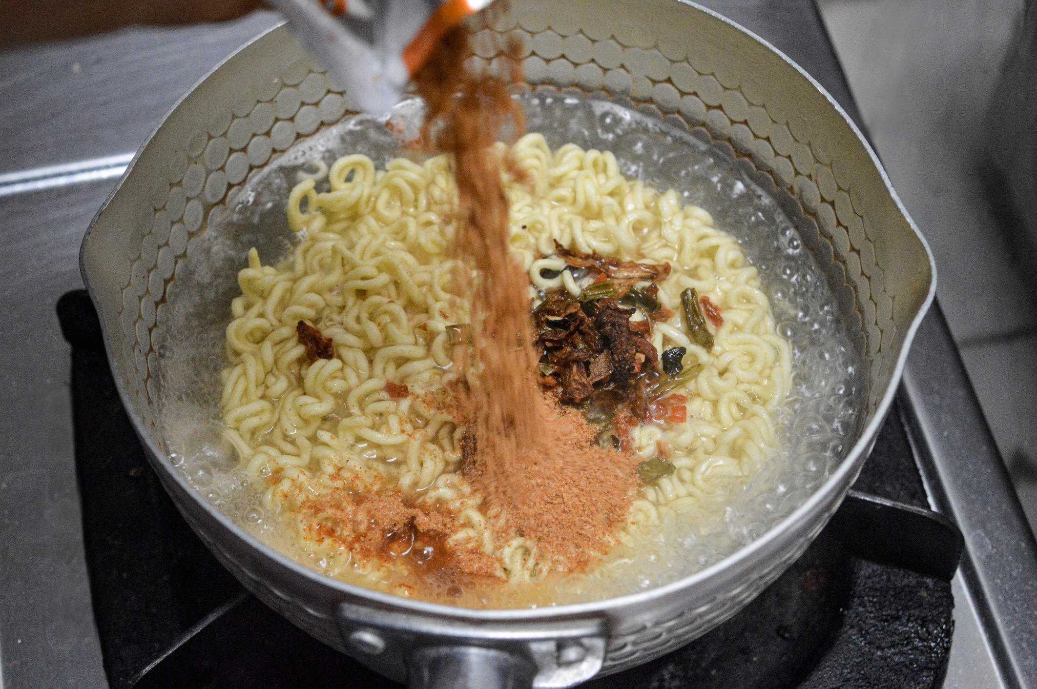 Instant noodles being cooked in a pot on a stove, with seasoning powder being added from a packet