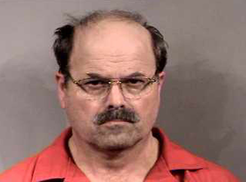 Man with glasses, mustache, and receding hairline wearing a plain shirt, looking directly at the camera