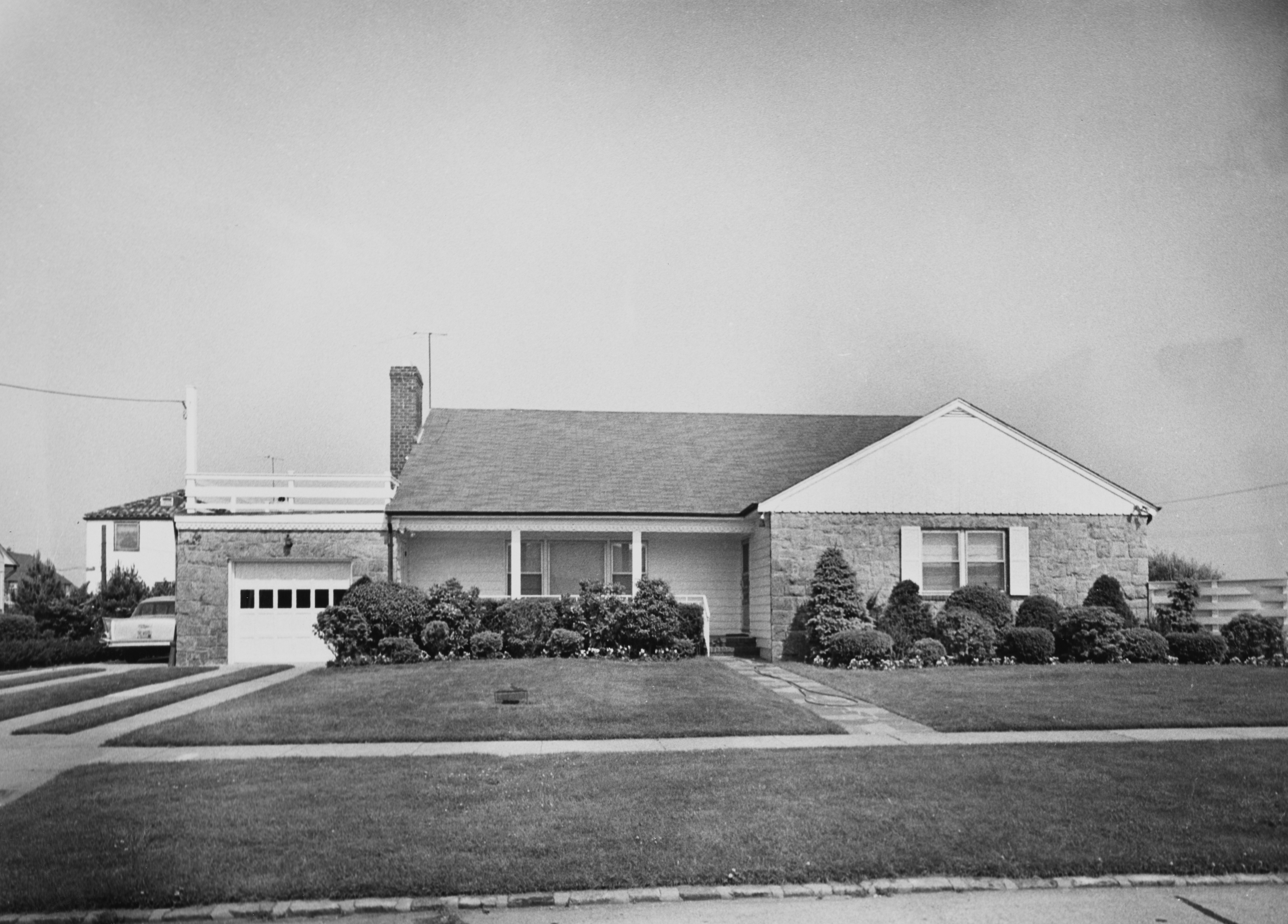 A black and white photograph of a single-story suburban house with a garage, well-kept lawn, and trimmed bushes. No people are in the image