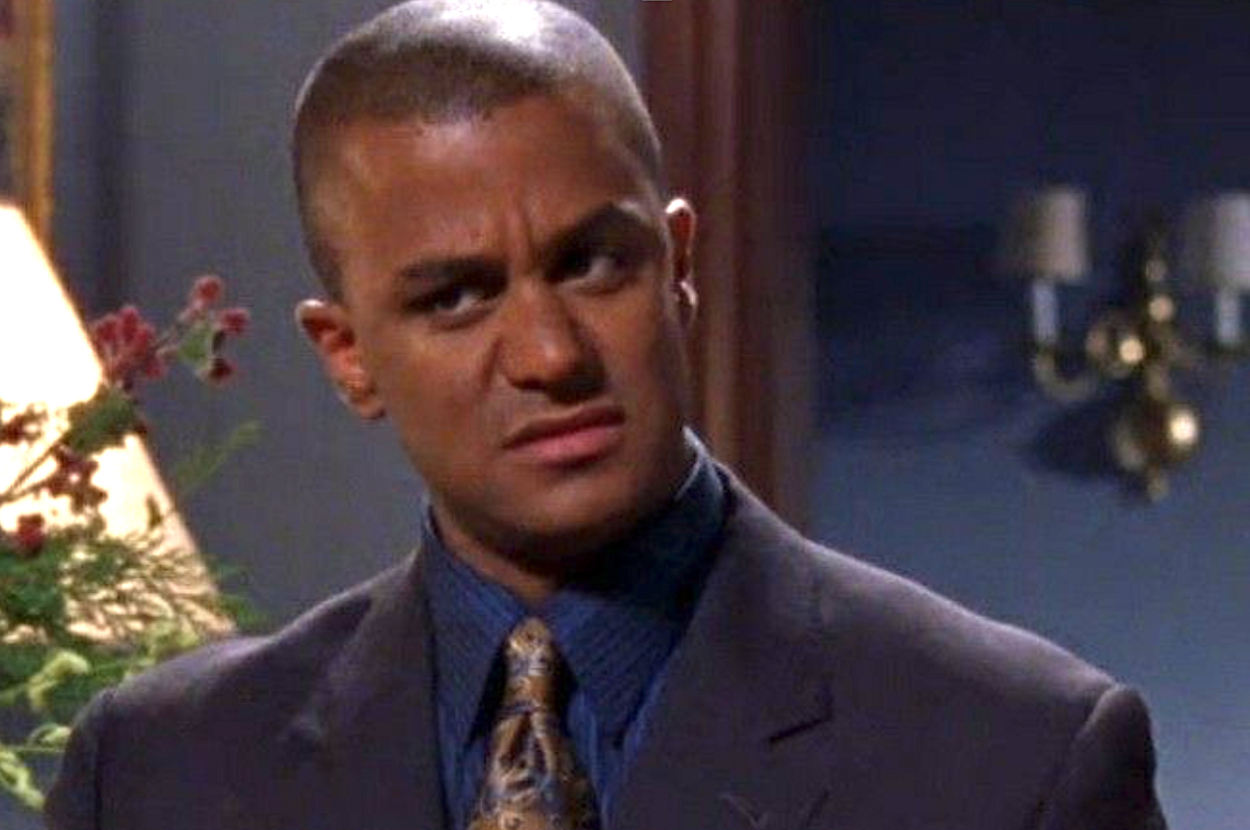 A man in a suit and tie looking puzzled, standing in a room with a decorative plant and wall light fixture in the background