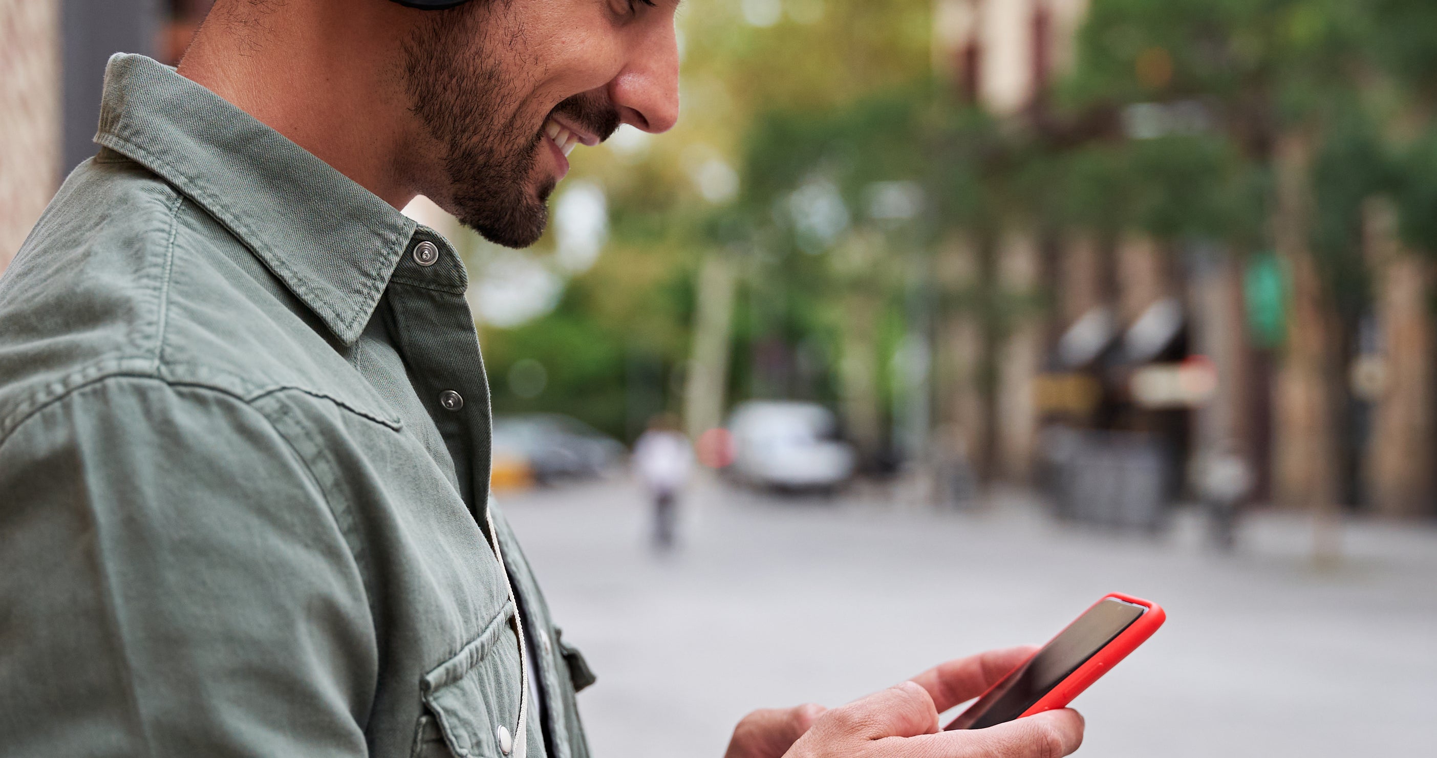A man wearing headphones is smiling while looking at his smartphone on a city street. He is leaning against a brick wall