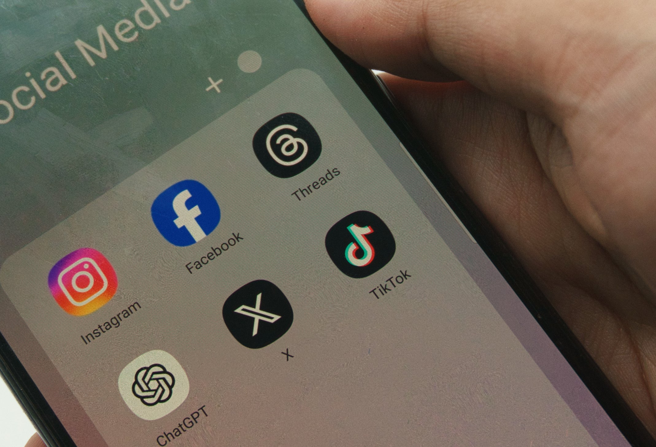 A hand holding a smartphone displaying a social media folder with Instagram, Facebook, Threads, X (formerly Twitter), TikTok, and ChatGPT apps