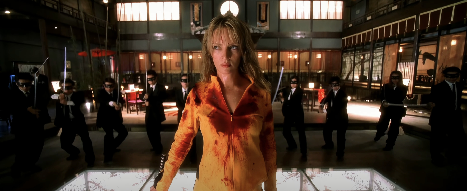 Uma Thurman, in a bloodstained yellow outfit, stands in a fighting pose surrounded by sword-wielding men in suits and masks in a Japanese-style interior