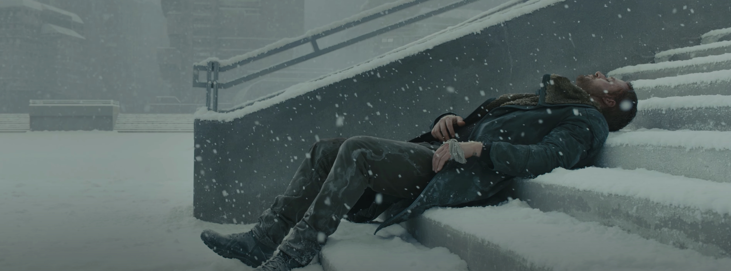 Man dressed warmly in a coat, lying on snowy steps outdoors in a storm