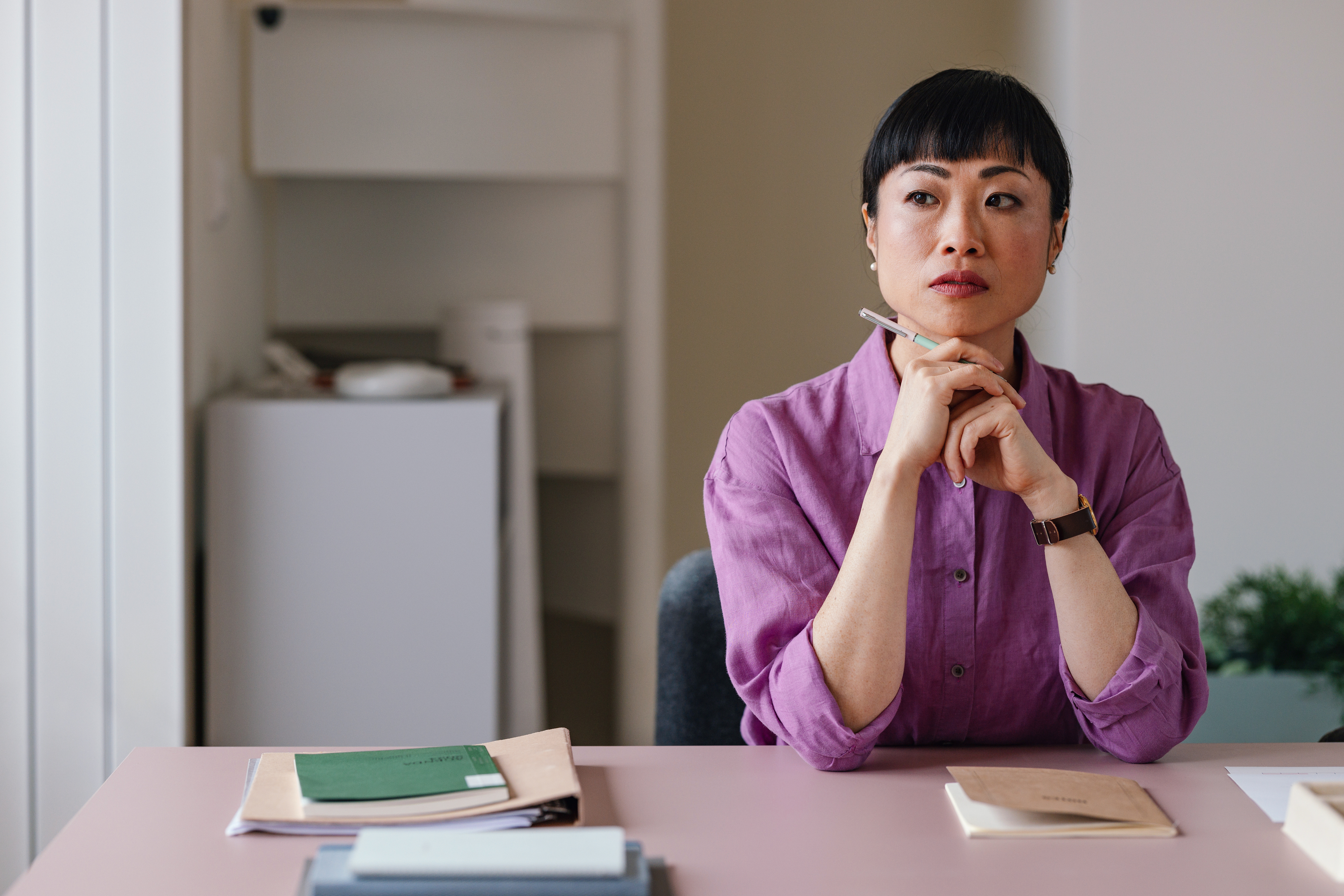 A person sitting at a desk, looking thoughtful, with a stack of papers and a pen in hand. The individual is wearing a collared shirt