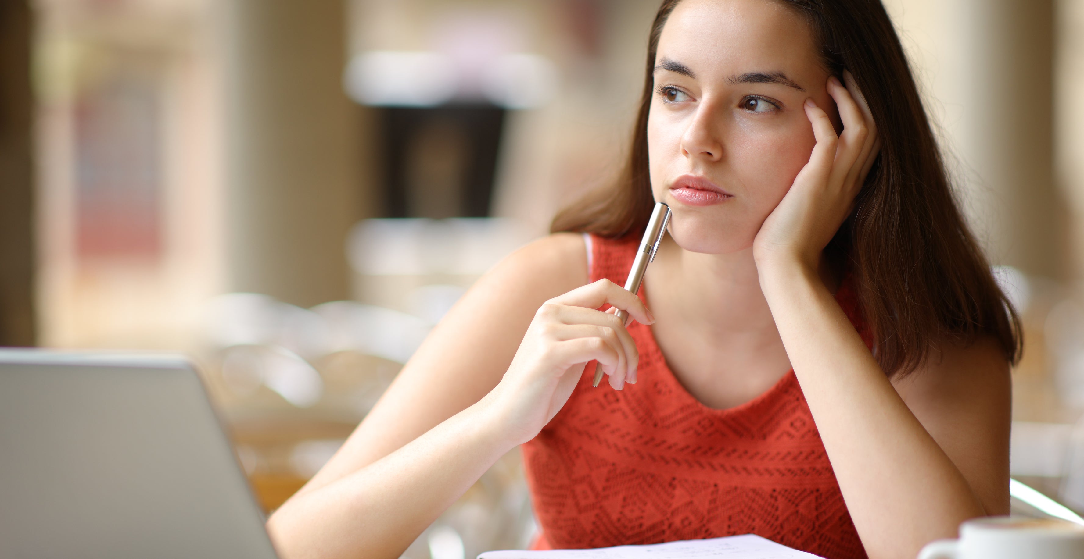 A woman in casual attire sits at a table, looking thoughtful, with a laptop and open notebook in front of her, holding a pen near her face