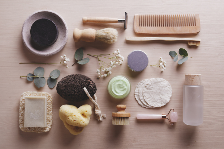 Various spa and grooming items including a bar of soap, exfoliating sponge, shaving brush, comb, facial cleanser, and dried flowers are arranged on a wooden surface