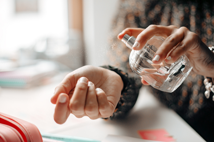 A person sprays perfume onto their wrist from a clear bottle, with various items in the blurred background